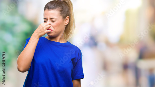 Young beautiful woman wearing casual blue t-shirt over isolated background smelling something stinky and disgusting, intolerable smell, holding breath with fingers on nose. Bad smells concept.
