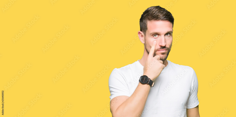 Handsome man wearing casual white t-shirt Pointing to the eye watching you gesture, suspicious expression Handsome man wearing casual white t-shirt Pointing to the eye watching you gesture, suspicious expression