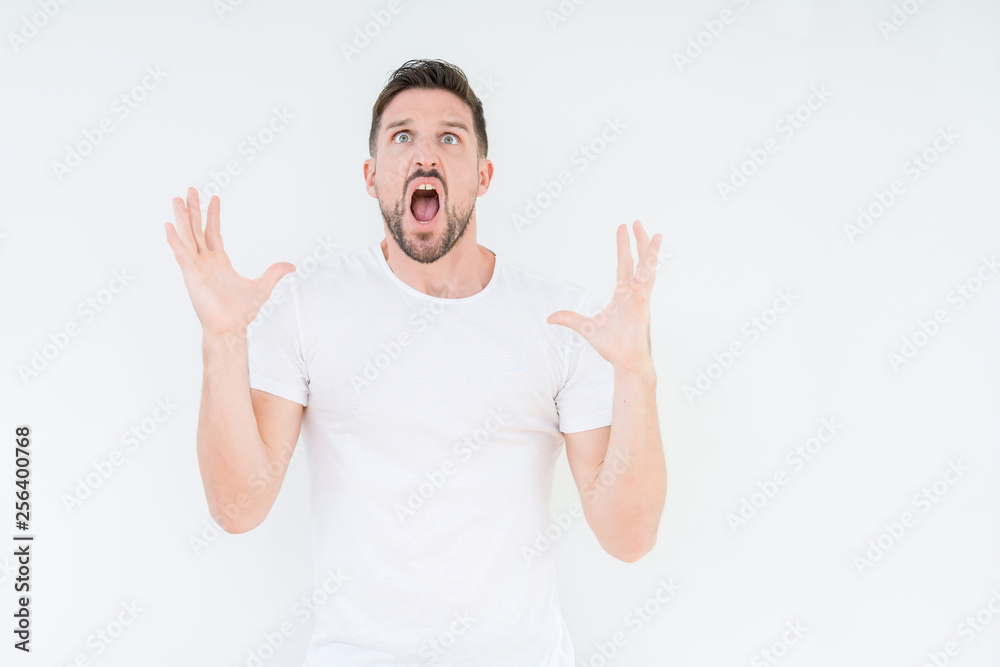 Fototapeta premium Young handsome man wearing casual white t-shirt over isolated background crazy and mad shouting and yelling with aggressive expression and arms raised. Frustration concept.