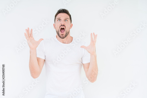 Young handsome man wearing casual white t-shirt over isolated background crazy and mad shouting and yelling with aggressive expression and arms raised. Frustration concept.