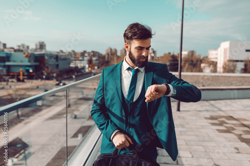 Young bearded Caucasian businessman in formal wear standing on rooftop and looking at wristwatch. In hand holding briefcase. Time is money.