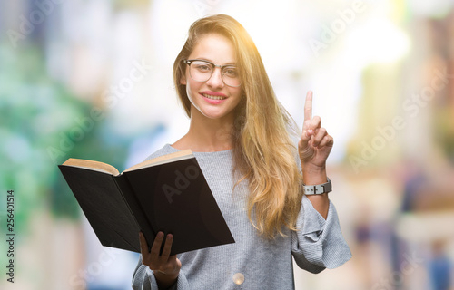 Young beautiful blonde woman reading a book over isolated background surprised with an idea or question pointing finger with happy face  number one