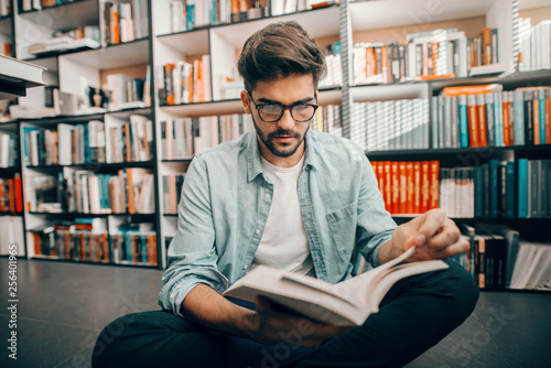 Mixed race male student sitting on library floor with legs crossed and reading book. In background books on shelves.