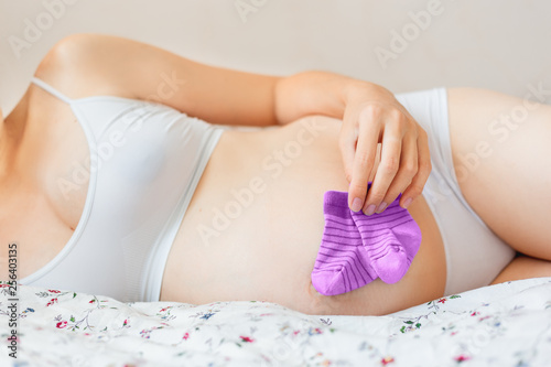 Pregnant woman in white underwear lying in bed with small baby's socks. Young woman expecting a baby girl. © Konstantin Aksenov