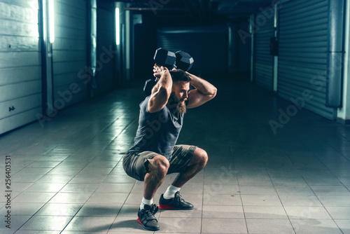 Side view of bearded Caucasian bodybuilder in sportswear doing exercises for backs while squatting in hallway at night.