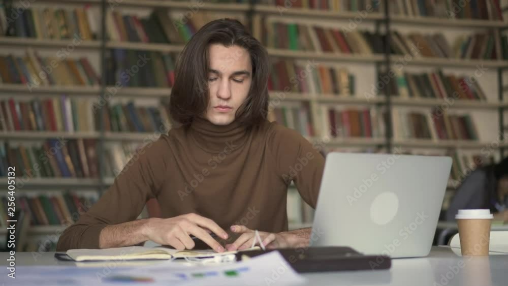 Pan shot of young man with long hair flipping the notebook sitting in library on the background of bookshelf. Young male student looking at the laptop preparing for exam. Pan shot of young man with long hair flipping the notebook sitting in library on the background of bookshelf. Young male student looking at the laptop preparing for exam.