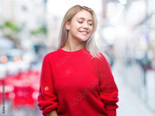 Young blonde woman wearing winter sweater over isolated background looking away to side with smile on face, natural expression. Laughing confident.