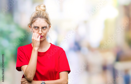 Young beautiful blonde woman wearing red t-shirt and glasses over isolated background looking stressed and nervous with hands on mouth biting nails. Anxiety problem.