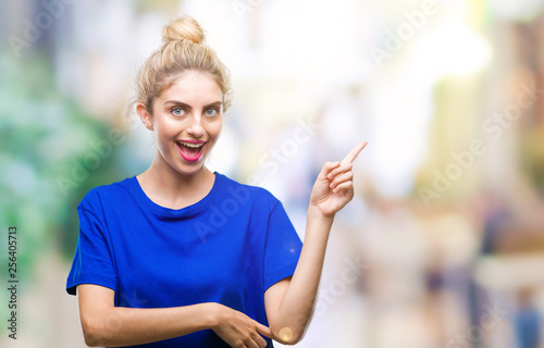 Young beautiful blonde and blue eyes woman wearing blue t-shirt over isolated background with a big smile on face, pointing with hand and finger to the side looking at the camera.