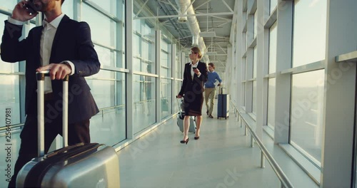 Business Caucasian people walking the airport passage with suitcases on wheels to the plane and speking on phone.