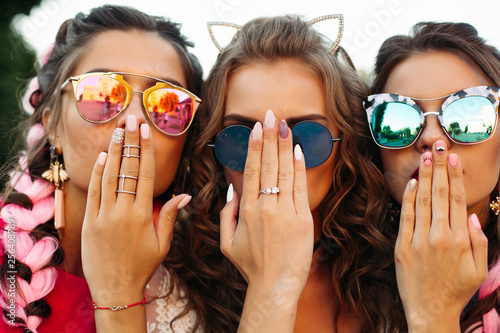 Close up of three young girls wearing in creative sunglasses with designed manicure, two lady covers mouth and one hiding face by hands. Happy friends posing and having fun together.