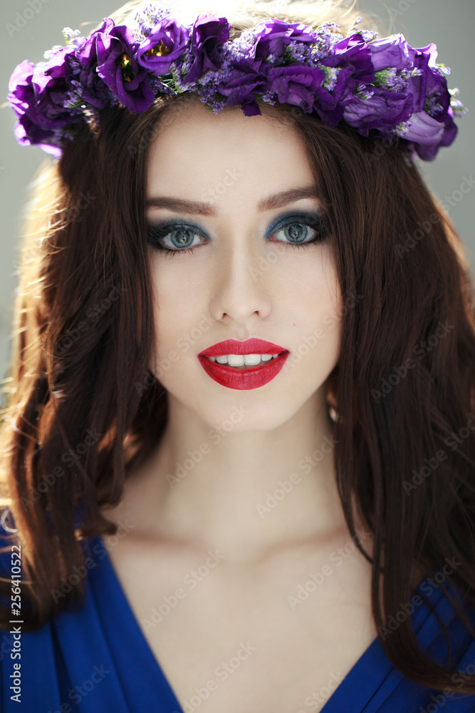 Naklejka premium Fashion Portrait of a beautiful brunette woman with amazing make up and coronet of purple flowers in her head, smiling at camera, outside background. Vertical view.