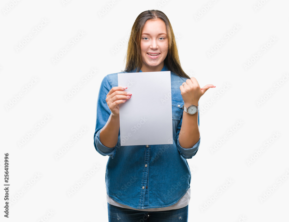 Young caucasian woman holding blank paper sheet over isolated background pointing and showing with thumb up to the side with happy face smiling Young caucasian woman holding blank paper sheet over isolated background pointing and showing with thumb up to the side with happy face smiling