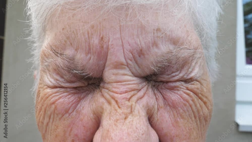 Close up wrinkled face of old grandmother looking into camera with a sad sight. Portrait of mature woman strongly screwing up her gray eyes. Sorrow facial expression of senior lady. Slow motion Close up wrinkled face of old grandmother looking into camera with a sad sight. Portrait of mature woman strongly screwing up her gray eyes. Sorrow facial expression of senior lady. Slow motion