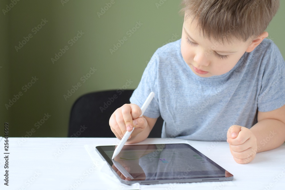 White caucasian smiling toddler boy drawing on tablet screen. Portrait of a happiness cute little boy with new tablet. Boy playing game on tablet on green blurred background and selective focus White caucasian smiling toddler boy drawing on tablet screen. Portrait of a happiness cute little boy with new tablet. Boy playing game on tablet on green blurred background and selective focus