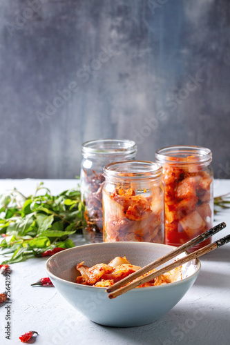 Korean traditional fermented appetizer kimchi cabbage and radish salad, hot spicy anchovies fish snack served in glass jars and bowl with Vietnamese oregano and chili peppers over grey blue table.