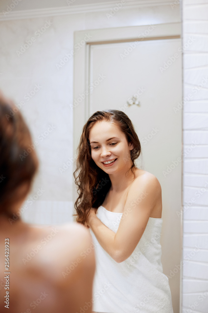 Beautiful young woman touching her wet hair with hands and smiling..Female looking of the mirror in bathroom. Care hair and skin. Girl uses protection moisturizing cream. Beautiful young woman touching her wet hair with hands and smiling..Female looking of the mirror in bathroom. Care hair and skin. Girl uses protection moisturizing cream.