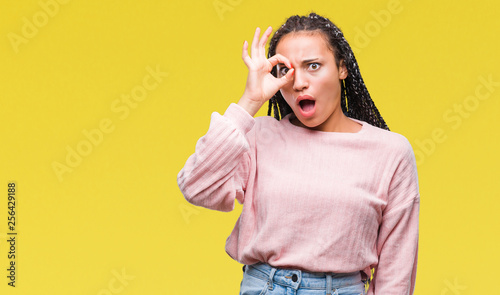 Young braided hair african american girl wearing sweater over isolated background doing ok gesture shocked with surprised face, eye looking through fingers. Unbelieving expression.