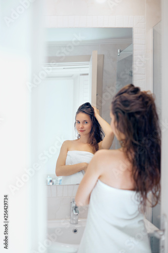 Hair and body care. Woman touching wet hair and smiling while looking in the mirror. Portrait of girl in bathroom applying conditioner and oil.Portrait of female uses protection moisturizing cream.