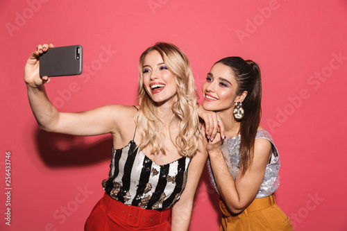 Fotografie Image of two attractive girls 20s in stylish outfit holding smartphone and takin