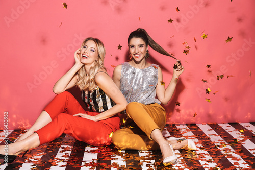 Portrait of two joyful women 20s in stylish outfit celebrating and sitting on floor with falling confetti