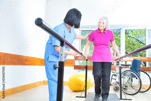 Asian nurse assisting aged senior woman training on the bars at rehab facility photo