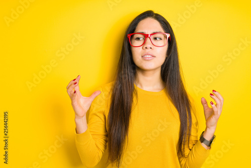 Beautiful brunette woman wearing red glasses over yellow isolated background crazy and mad shouting and yelling with aggressive expression and arms raised. Frustration concept.
