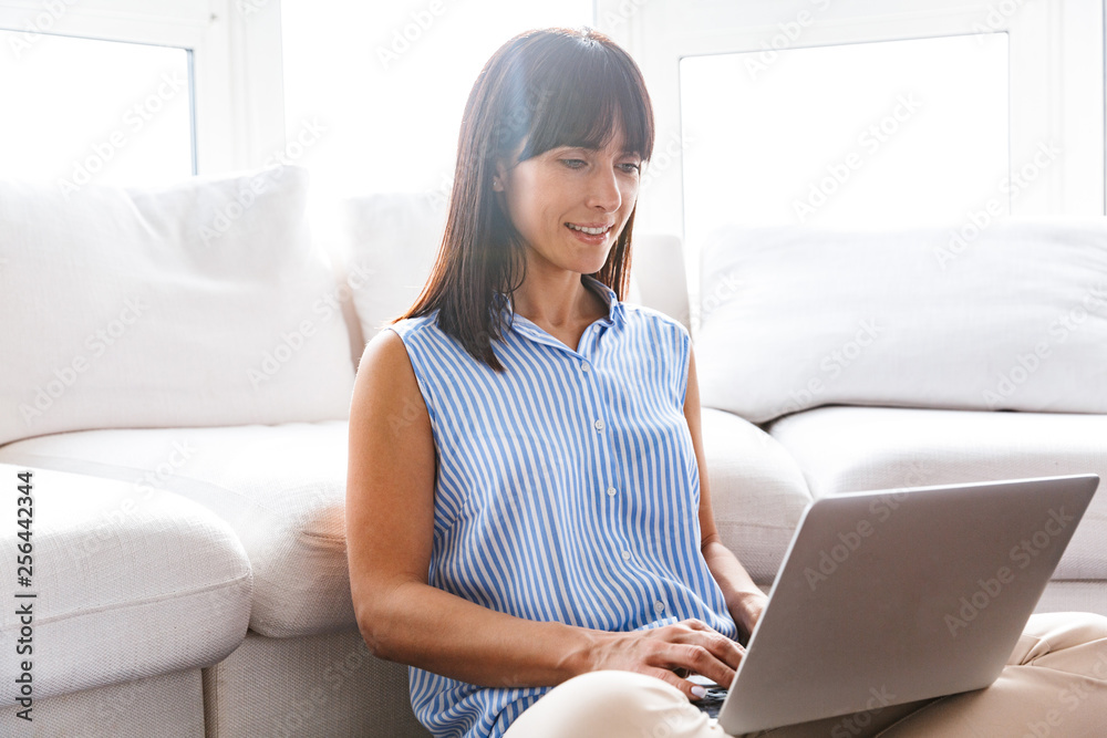 Naklejka premium Portrait of attractive woman 40s using silver laptop while sitting on couch in bright apartment