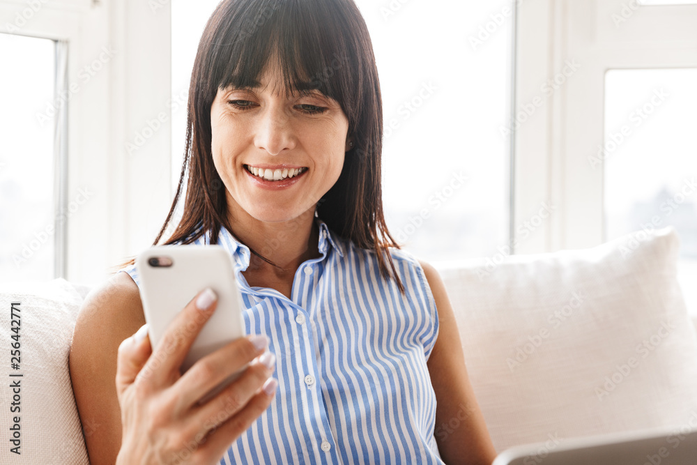 Portrait of happy woman 40s using cell phone and laptop while sitting on couch in bright apartment Portrait of happy woman 40s using cell phone and laptop while sitting on couch in bright apartment