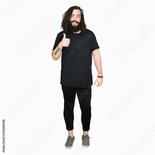 Young man with long hair and beard wearing heavy metal black outfit doing happy thumbs up gesture with hand. Approving expression looking at the camera with showing success.