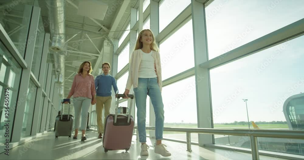 Caucasian teen cute and pretty girl walking the airport corridor with a suitcase on the wheels to the plane and her parents following. Caucasian teen cute and pretty girl walking the airport corridor with a suitcase on the wheels to the plane and her parents following.