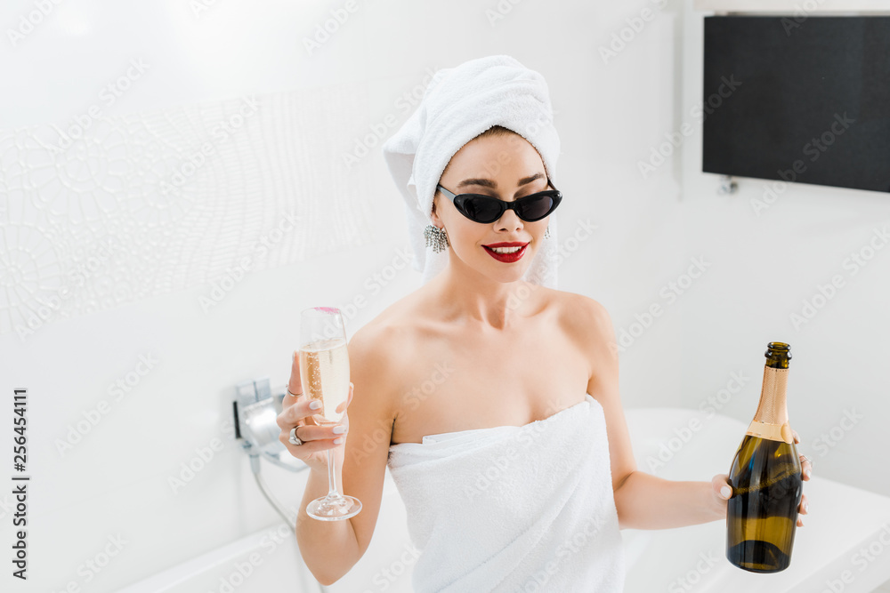 attractive and happy woman in sunglasses and towels holding champagne glass and bottle attractive and happy woman in sunglasses and towels holding champagne glass and bottle