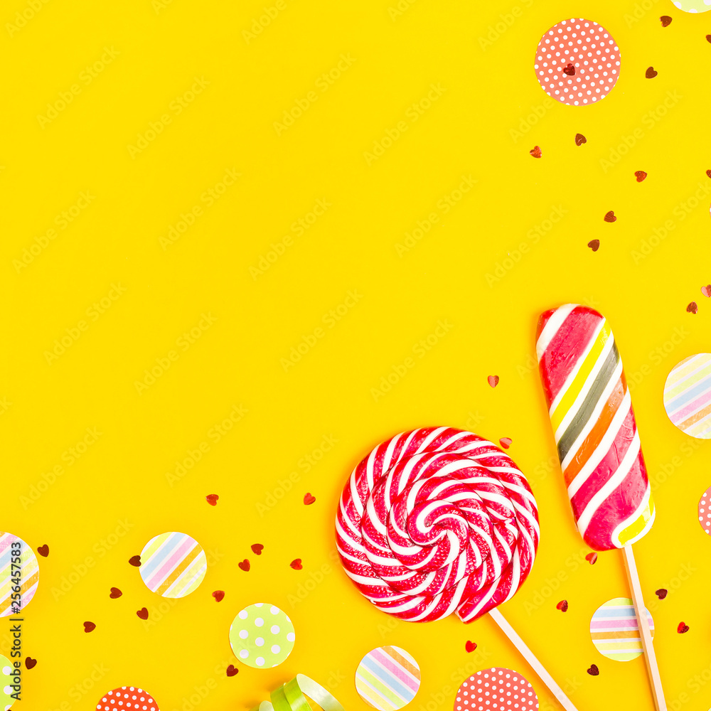 Two multicolored candies among paper circles of confetti, glitter and festive ribbons on a bright yellow background. Top view, flat lay, copy space Two multicolored candies among paper circles of confetti, glitter and festive ribbons on a bright yellow background. Top view, flat lay, copy space