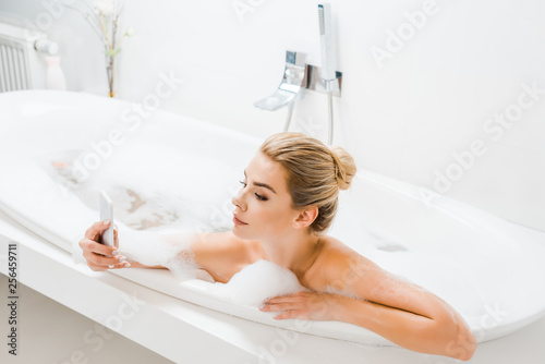 beautiful and blonde woman taking bath with foam and using smartphone in bathroom
