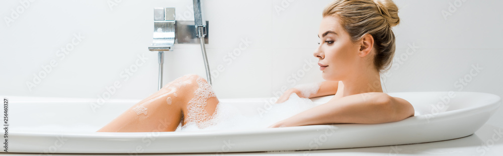 panoramic shot of beautiful and blonde woman taking bath with foam and looking away in bathroom panoramic shot of beautiful and blonde woman taking bath with foam and looking away in bathroom