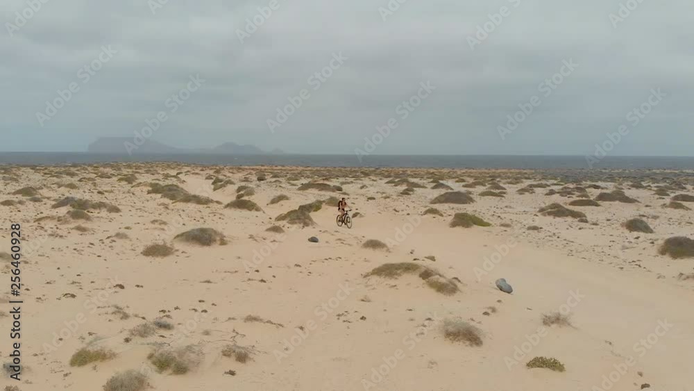 Woman riding bicycle on a sandy beach. Female mountain biking on a cloudy day over the sea in a desert path over volcanic island of La Graciosa Woman riding bicycle on a sandy beach. Female mountain biking on a cloudy day over the sea in a desert path over volcanic island of La Graciosa