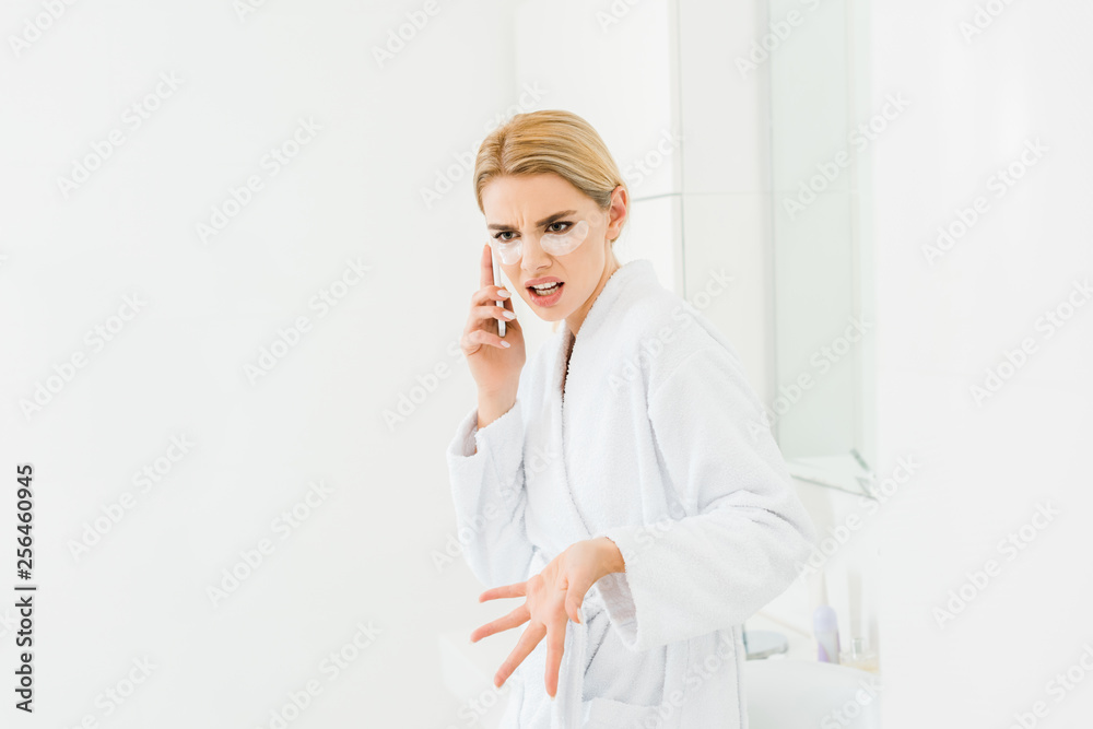 beautiful and irritated woman in white bathrobe with eye patches on face talking on smartphone beautiful and irritated woman in white bathrobe with eye patches on face talking on smartphone