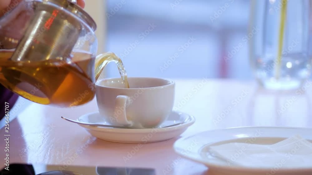 Female hand pours brown tea from a transparent kettle in a white cup on a table in a cafe Female hand pours brown tea from a transparent kettle in a white cup on a table in a cafe