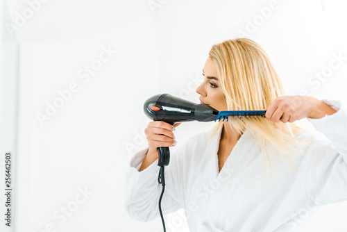 beautiful and smiling woman in white bathrobe using hairdryer and comb