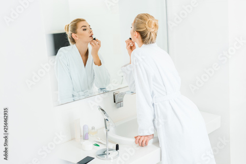 selective focus of beautiful and blonde woman in white bathrobe applying lipstick in bathroom