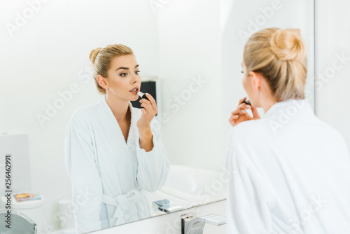 selective focus of woman in white bathrobe applying lipstick and looking at mirror in bathroom