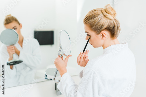selective focus of woman in white bathrobe applying eyeshadow with cosmetic brush