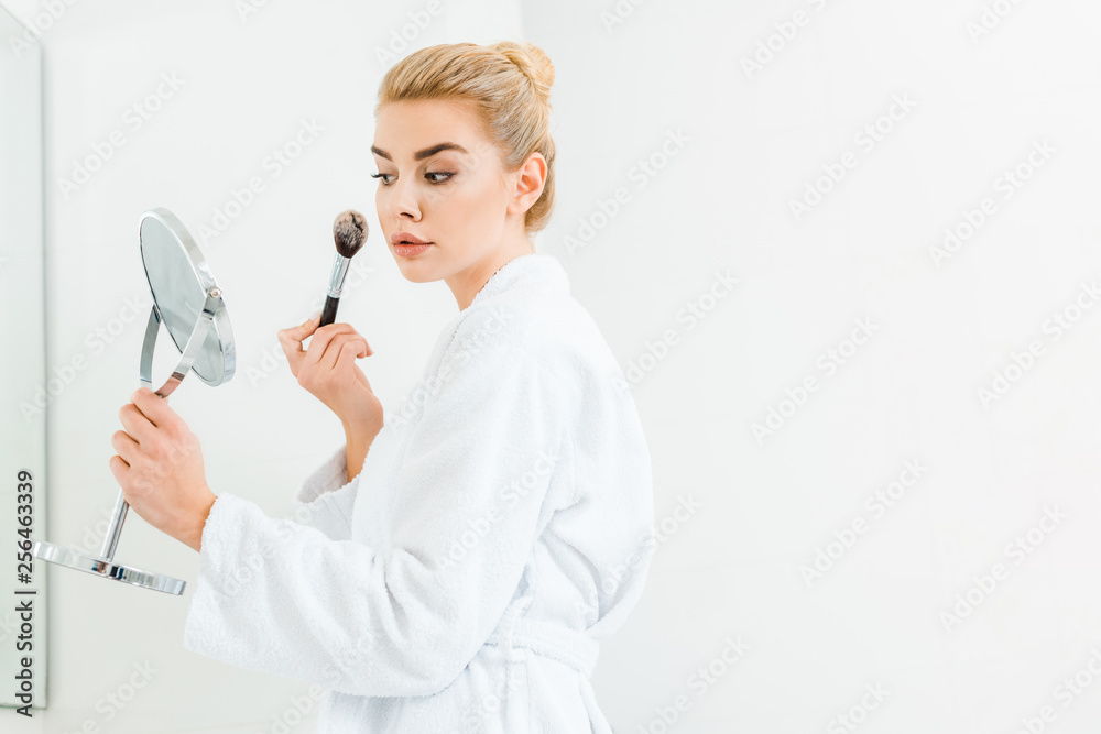 beautiful and blonde woman in white bathrobe using cosmetic brush in bathroom beautiful and blonde woman in white bathrobe using cosmetic brush in bathroom