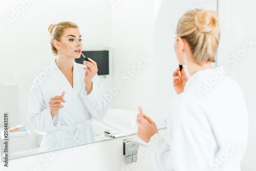 selective focus of woman in white bathrobe applying lip gloss in bathroom