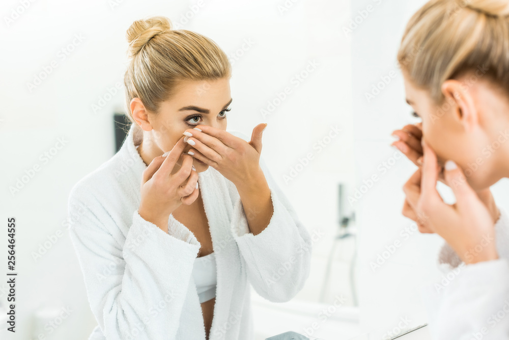 selective focus of attractive and blonde woman in white bathrobe attaching contact lens in bathroom selective focus of attractive and blonde woman in white bathrobe attaching contact lens in bathroom