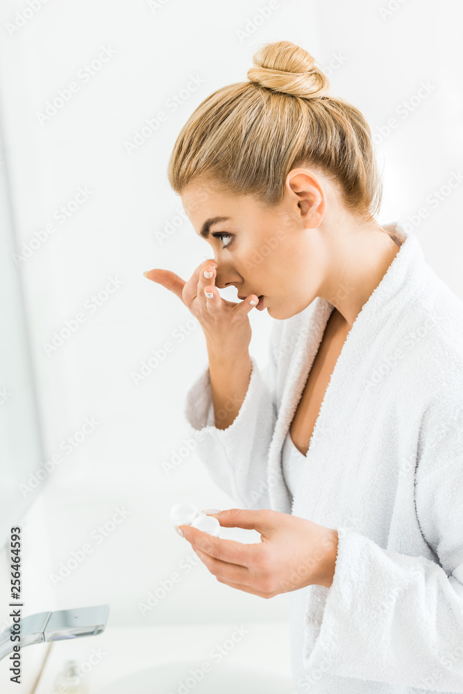 beautiful and blonde woman in white bathrobe attaching contact lens in bathroom beautiful and blonde woman in white bathrobe attaching contact lens in bathroom