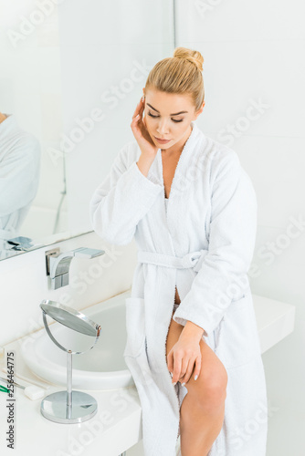 attractive and blonde woman in white bathrobe looking at mirror in bathroom