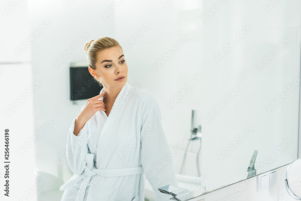 beautiful and blonde woman in white bathrobe looking at mirror in bathroom beautiful and blonde woman in white bathrobe looking at mirror in bathroom