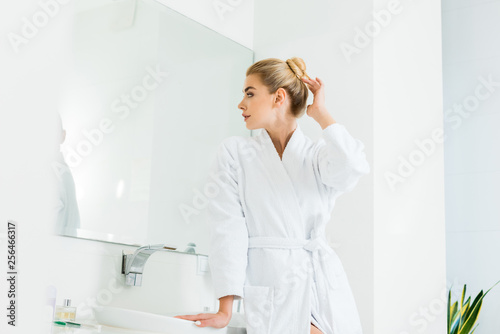 attractive and blonde woman in white bathrobe looking at mirror in bathroom