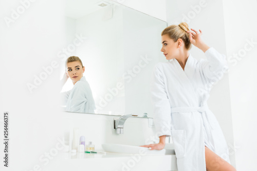 selective focus of attractive and blonde woman in white bathrobe looking at mirror in bathroom